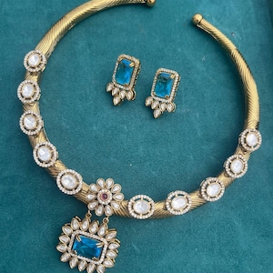 Puede incluir: Un collar dorado y un conjunto de pendientes a juego. El collar presenta una banda texturizada con detalles circulares de piedras transparentes. El colgante tiene una gran piedra azul rectangular. Los pendientes tienen piedras azules rectangulares.