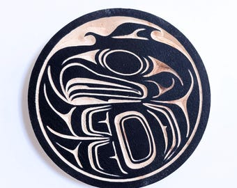 May include: Black and white circular wooden coaster with a carved design of a stylized orca whale. The design is in the style of Northwest Coast Indigenous art.