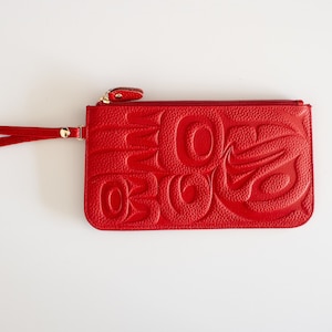 Puede incluir: Un clutch de cuero rojo con cierre de cremallera y una correa de muñeca. El clutch presenta un diseño en relieve de un animal estilizado, posiblemente un pájaro o un pez.