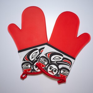 Könnte beinhalten: Ein Paar rote Silikon-Ofenhandschuhe mit einem weißen und schwarzen Design, das einen stilisierten Raben und andere Vögel zeigt. Das Design erinnert an die indigene Kunst der Nordwestküste.