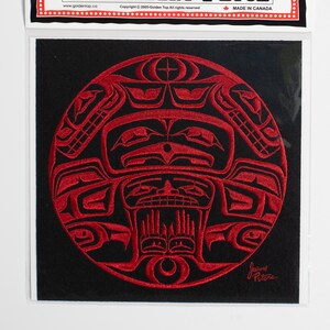 Könnte beinhalten: Roter gestickter Aufnäher mit einem kreisförmigen Design, das ein stilisiertes Gesicht mit geometrischen Mustern zeigt. Der Aufnäher befindet sich auf einem schwarzen Hintergrund und trägt den Text "Golden Top Embroidery Patch" und "Hergestellt in Kanada" auf der Verpackung.