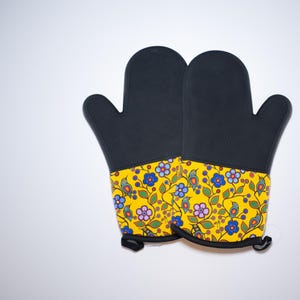 Könnte beinhalten: Ein Paar schwarze Ofenhandschuhe mit gelbem Blumenmuster. Die Handschuhe haben eine Aufhängeschlaufe.