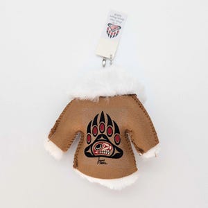 May include: A brown miniature jacket with white faux fur trim and a Native American-style bear paw design in red and black. The jacket has a tag attached with text and a Native American design.