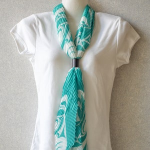 May include: A white t-shirt with a teal and white patterned scarf tied around the neck. The scarf has a black ring holding it in place.