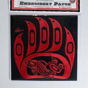 Könnte beinhalten: Roter gestickter Aufnäher mit einer Bärenpfote, in der sich ein stilisierter Lachs befindet. Der Aufnäher ist auf schwarzem Hintergrund und trägt oben den Text "Golden Top Embroidery Patch".