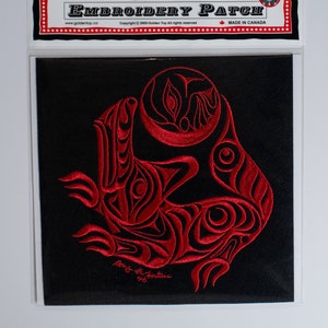 以下が含まれることがあります： 黒い正方形の刺繍パッチ。赤い、様式化された生き物のデザインが施されています。パッチは透明なプラスチック製のスリーブに入っています。上部には「EMBROIDERY PATCH」の文字と「MADE IN CANADA」の文字が見えます。