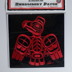 Può includere: Una toppa ricamata con un disegno di aquila rossa stilizzata su sfondo nero. La toppa è confezionata in una custodia di plastica trasparente con un'intestazione che recita "EMBROIDERY PATCH" e "MADE IN CANADA". Il logo Golden Top è visibile.