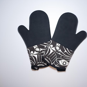 Op de afbeelding: Een paar zwarte ovenwanten met een wit en zwart patroon. Het patroon bevat gestileerde dierfiguren.