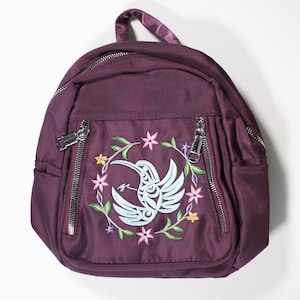 Pode incluir: Uma mochila cor de ameixa com um design bordado central. O design apresenta um beija-flor estilizado em azul claro e branco, rodeado por uma coroa floral com flores rosa, amarelas e roxas. A mochila tem dois bolsos com zíper e uma alça superior.