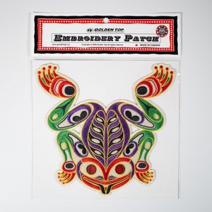 Könnte beinhalten: Ein gestickter Aufnäher mit einem farbenfrohen Frosch in einem stilisierten Design. Der Frosch ist grün, lila, rot, gelb und schwarz. Der Aufnäher befindet sich auf einem weißen Hintergrund mit einer Karte, auf der "Golden Top Embroidery Patch" und "Made in Canada" steht.
