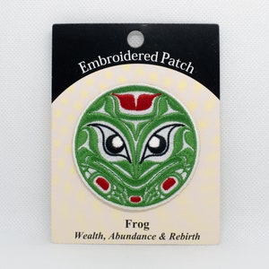 May include: Embroidered patch with a green frog design. The frog has large eyes and a red mouth. The patch is on a white card with the words "Embroidered Patch" and "Frog, Wealth, Abundance & Rebirth".