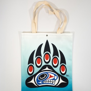 Könnte beinhalten: Eine weiße Canvas-Tragetasche mit einem blauen und schwarzen Bärenpfoten-Design. Die Pfote hat rote Kreise für die Krallen und ein stilisiertes Gesicht in der Mitte. Die Tasche hat cremefarbene Henkel.