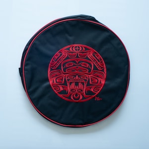 May include: A black round drum bag with a red embroidered design of a stylised face with a crown. The bag has a red trim around the edge.