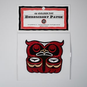 Könnte beinhalten: Gestickter Aufnäher mit einem stilisierten Eulen-Design in Rot, Schwarz und Gold. Der Aufnäher befindet sich in einer transparenten Kunststoffhülle mit der Aufschrift "EMBROIDERY PATCH" und dem Golden Top-Logo.