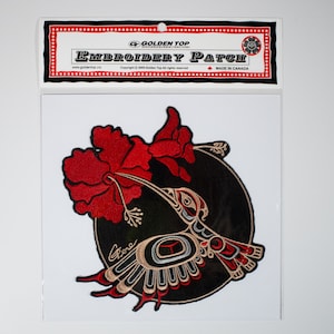 Puede incluir: Un parche bordado con un colibrí estilizado con detalles rojos, negros y dorados, bebiendo néctar de una flor de hibisco roja. El parche es circular y tiene un fondo negro. El texto "Golden Top Embroidery Patch" está impreso en el embalaje.