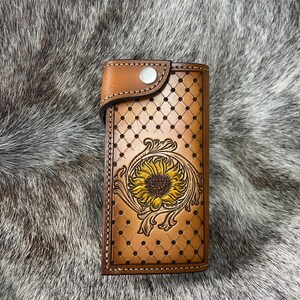 May include: A brown leather wallet with a sunflower design. The wallet features a detailed embossed sunflower with yellow petals and a brown center. The background has a diamond pattern. The wallet is secured with a snap closure.
