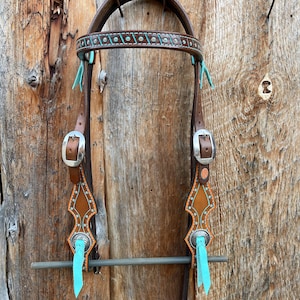 May include: A brown leather horse bridle with turquoise stitching and accents. The bridle is hanging on a wooden wall with nails.