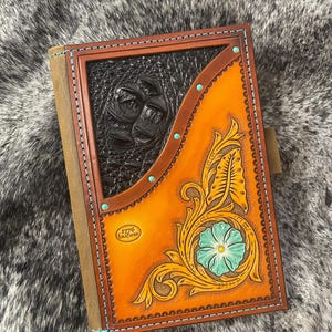 May include: A brown leather journal with a tooled design featuring a turquoise flower and leaves. The journal has a black leather panel with a tooled design and the text "1776 Leather" in a circle.