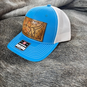 May include: A blue trucker hat with a white mesh back and a curved brim. The hat features a brown leather patch with a floral and bull skull design. The Richardson 112 hat is adjustable.