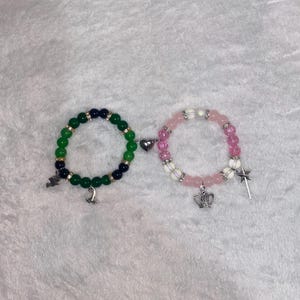 Könnte beinhalten: Zwei Armbänder mit Perlen, verbunden durch einen Herzanhänger. Ein Armband hat grüne und marinefarbene Perlen mit einem Anhänger, das andere rosa und weiße Perlen mit einem Kronen- und Sternanhänger. Auf weißer, strukturierter Oberfläche.