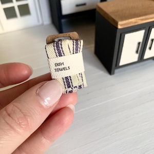 May include: A miniature set of dish towels with a purple and white striped pattern. The towels are folded and tied with a brown ribbon. The label reads "Dish Towels".