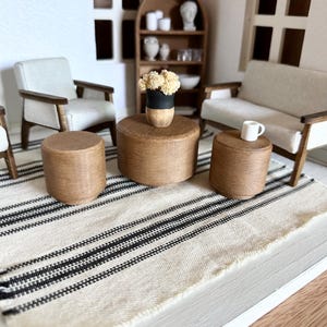 May include: Miniature living room scene with a neutral color palette. Features a sofa, two armchairs, and two small round tables. A black and white striped rug and a small vase with flowers are also present. The furniture has wooden frames.
