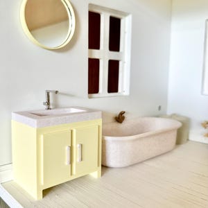 May include: A miniature bathroom scene featuring a yellow vanity with a speckled countertop and a silver faucet. A round gold mirror hangs on the wall, and a bathtub is in the background. The room has a window and light wood flooring.