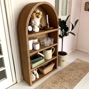 May include: A wooden arched shelf unit with five shelves, featuring decorative items such as a white bust, a clock, glass jars, glasses, a pitcher, books, a woven basket, and a pair of white swans. The shelf unit is against a white wall with a brown rug in front of it.