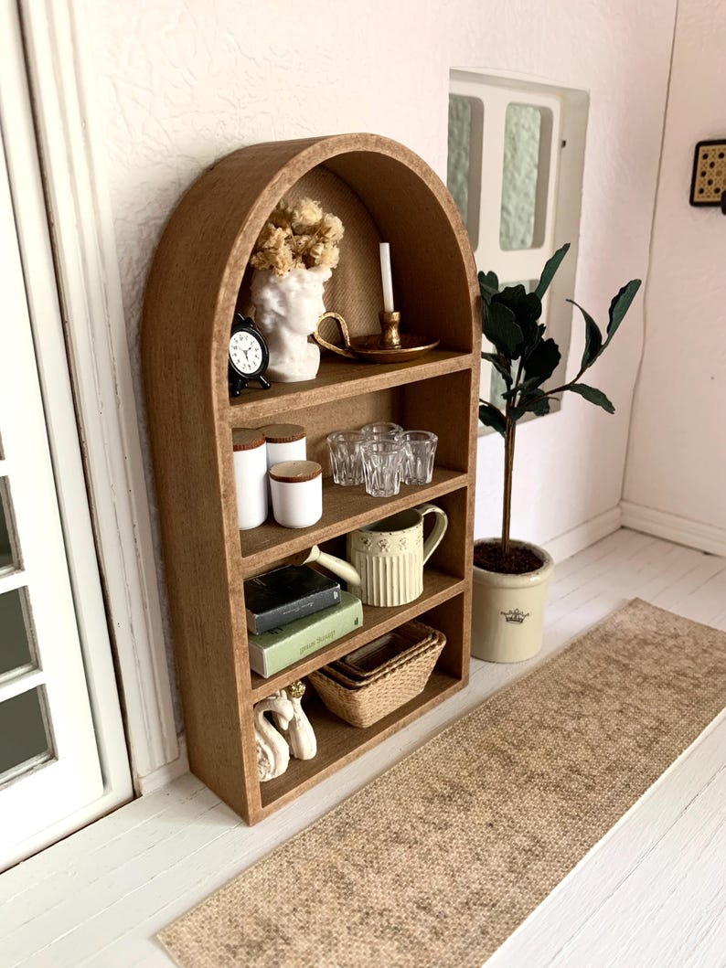 May include: A wooden arched shelf unit with five shelves, featuring decorative items such as a white bust, a clock, glass jars, glasses, a pitcher, books, a woven basket, and a pair of white swans. The shelf unit is against a white wall with a brown rug in front of it.