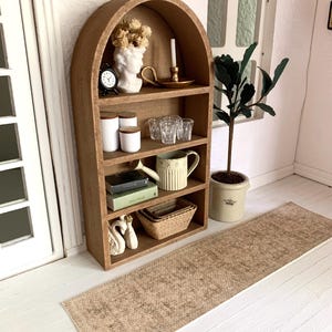 May include: A wooden arched shelf unit with four shelves, filled with decorative items, including a bust, a clock, glasses, books, and a basket. The shelf is against a white wall with a brown rug in front of it.