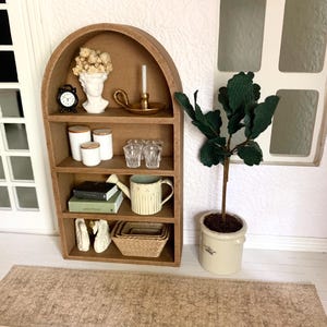 May include: A miniature wooden arched shelf with four shelves, featuring a white bust, a small clock, a candle holder, glass cups, a watering can, books, white swans, and a woven basket. The shelf is against a white wall with a window and a potted green plant.
