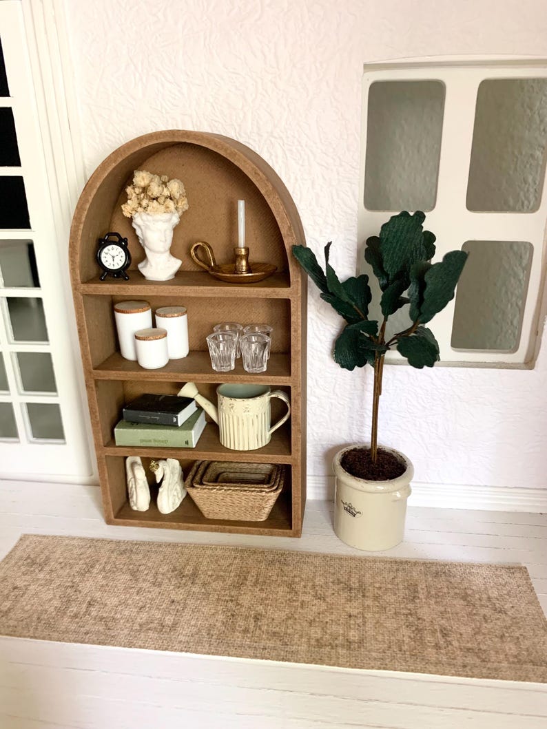 May include: A miniature wooden arched shelf with four shelves, featuring a white bust, a small clock, a candle holder, glass cups, a watering can, books, white swans, and a woven basket. The shelf is against a white wall with a window and a potted green plant.