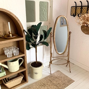 May include: A miniature dollhouse room with a gold-framed oval mirror, a potted green plant, a wooden shelf with a watering can, glasses, and a candle, and a woven rug on the floor.