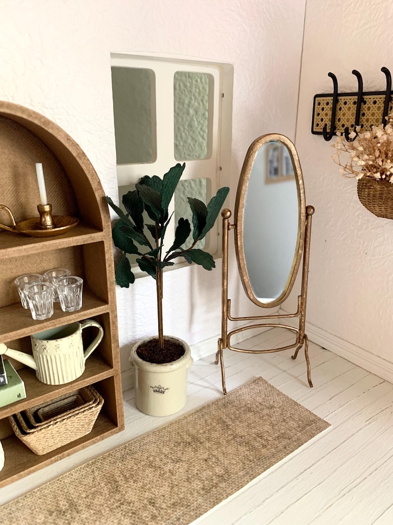 May include: A miniature dollhouse room with a gold-framed oval mirror, a potted green plant, a wooden shelf with a watering can, glasses, and a candle, and a woven rug on the floor.