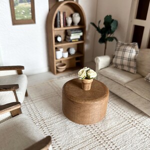 Op de afbeelding: Miniatuur poppenhuis woonkamer sc&egrave;ne met een ronde houten salontafel, fauteuils, een bank en een boekenplank. Een kleine pot met witte bloemen staat op de tafel. De kamer heeft een neutraal kleurenpalet met een patroonkleed.