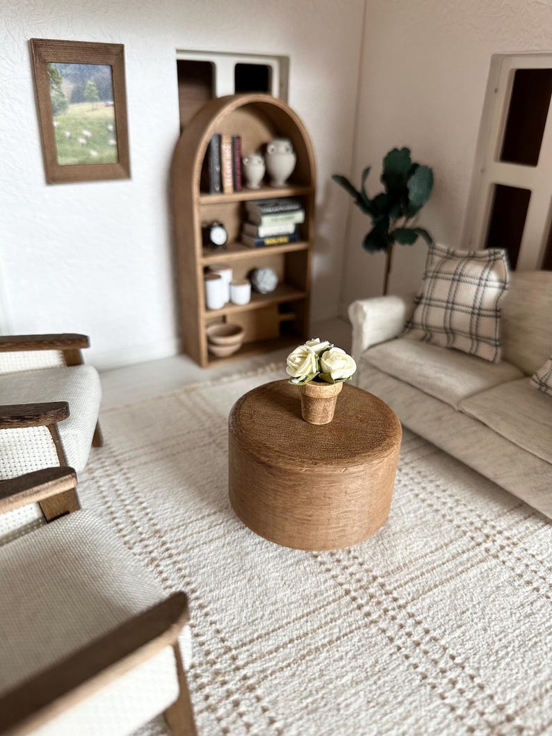 Op de afbeelding: Miniatuur poppenhuis woonkamer sc&egrave;ne met een ronde houten salontafel, fauteuils, een bank en een boekenplank. Een kleine pot met witte bloemen staat op de tafel. De kamer heeft een neutraal kleurenpalet met een patroonkleed.