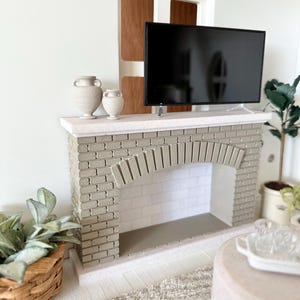 May include: Miniature fireplace with a gray brick facade, a black flat-screen TV, and two decorative vases. The fireplace has an arched opening and is set against a white wall. A small plant and a woven basket are in the foreground.