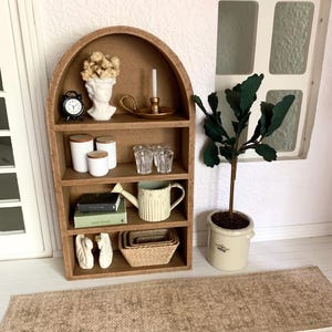 May include: A miniature wooden arched shelf unit with five shelves, featuring a variety of decorative items, including a white bust, a small clock, a candle holder, a watering can, books, and a potted plant. The shelf unit is set on a brown rug.
