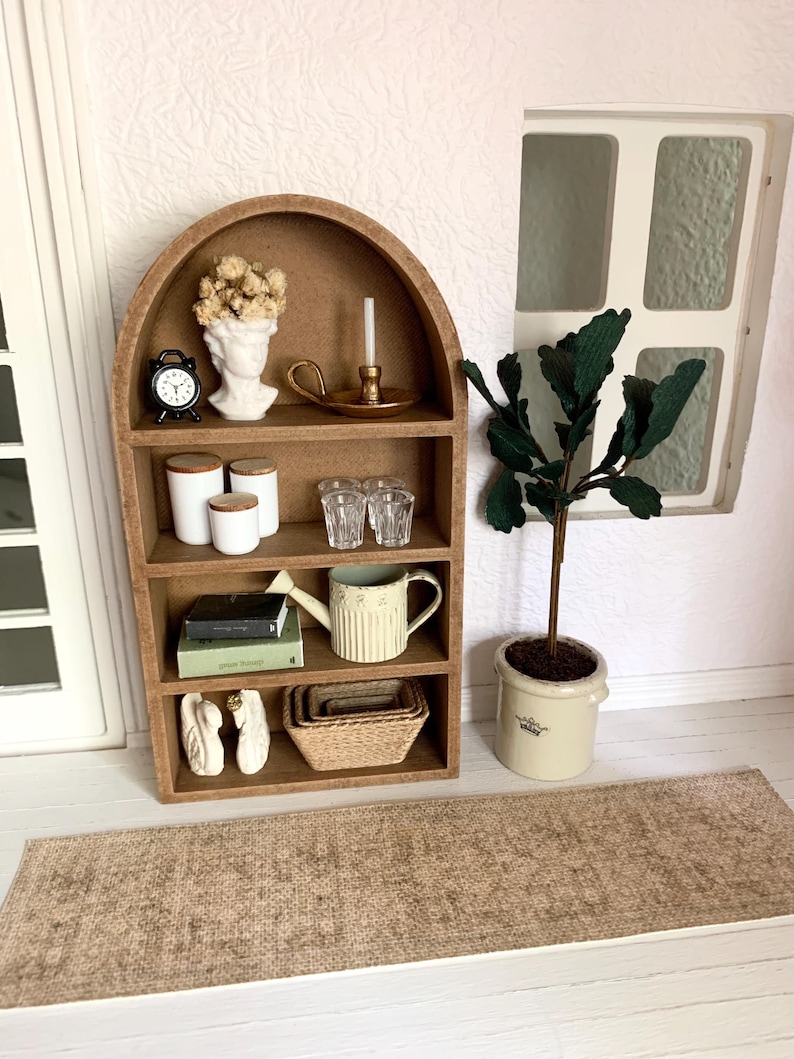 May include: A miniature wooden arched shelf unit with five shelves, featuring a variety of decorative items, including a white bust, a small clock, a candle holder, a watering can, books, and a potted plant. The shelf unit is set on a brown rug.