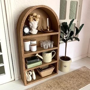 May include: A miniature wooden arched shelf with five shelves, featuring decorative items such as a white bust, a small clock, a candle holder, glass cups, a watering can, books, and a woven basket. The shelf is set against a white wall with a brown rug in the foreground.
