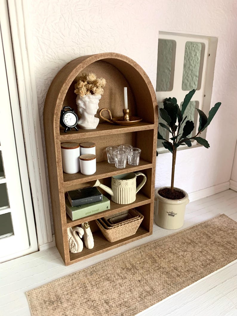 May include: A miniature wooden arched shelf with five shelves, featuring decorative items such as a white bust, a small clock, a candle holder, glass cups, a watering can, books, and a woven basket. The shelf is set against a white wall with a brown rug in the foreground.
