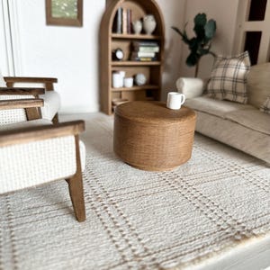 Op de afbeelding: Miniatuur woonkamer sc&egrave;ne met een lichtbruine houten salontafel, een witte mok en een beige geruit tapijt. Op de achtergrond een lichtgekleurde bank met een geruit kussen en een houten boekenkast. Een ingelijste foto hangt aan de muur.