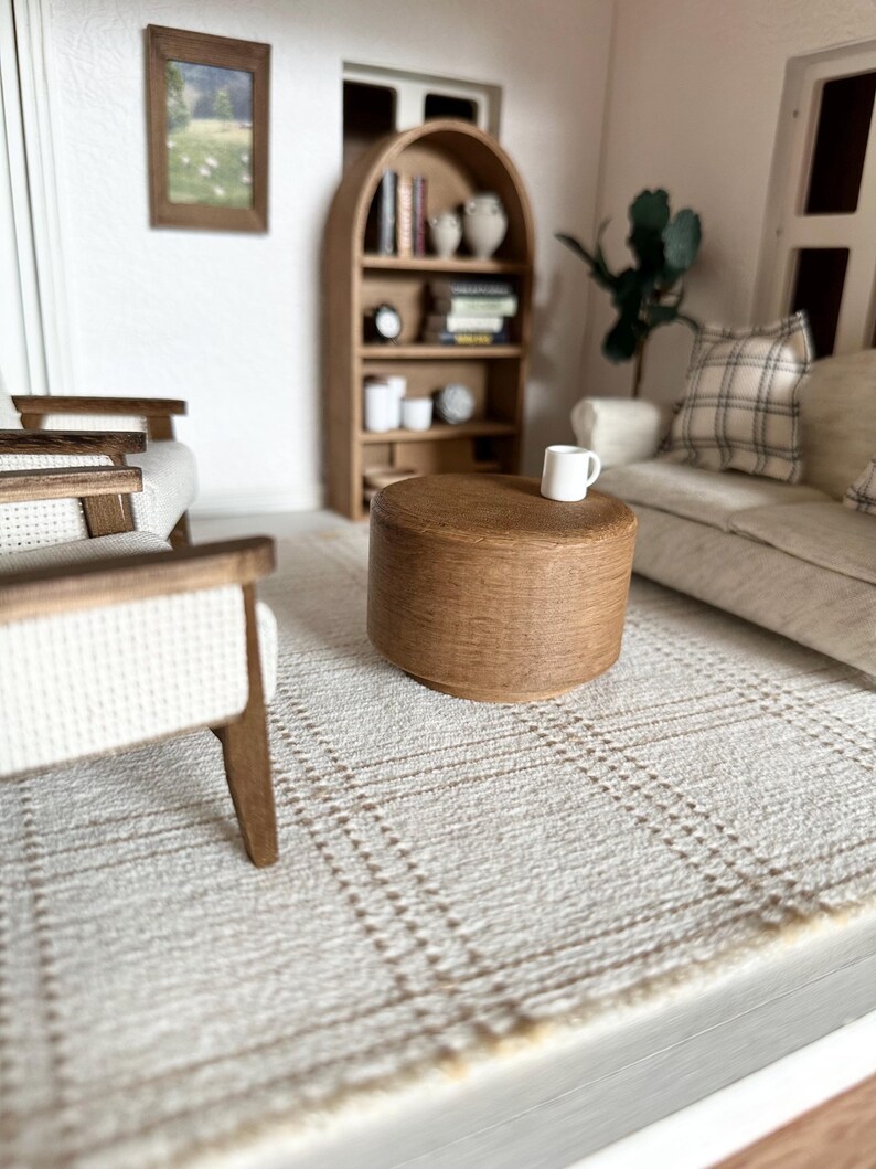 Op de afbeelding: Miniatuur woonkamer sc&egrave;ne met een lichtbruine houten salontafel, een witte mok en een beige geruit tapijt. Op de achtergrond een lichtgekleurde bank met een geruit kussen en een houten boekenkast. Een ingelijste foto hangt aan de muur.