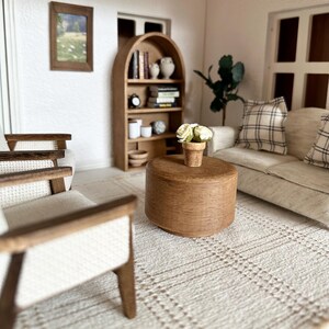 Op de afbeelding: Miniatuur poppenhuis woonkamer met een beige bank, geruite kussens en een ronde houten salontafel. Op de tafel staat een kleine pot met witte bloemen. Een houten boekenkast en ingelijste kunstwerken zijn ook zichtbaar.