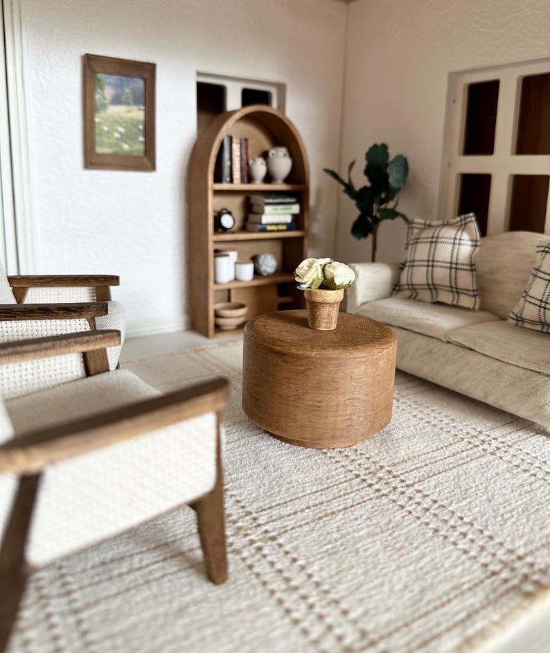 Op de afbeelding: Miniatuur poppenhuis woonkamer met een beige bank, geruite kussens en een ronde houten salontafel. Op de tafel staat een kleine pot met witte bloemen. Een houten boekenkast en ingelijste kunstwerken zijn ook zichtbaar.