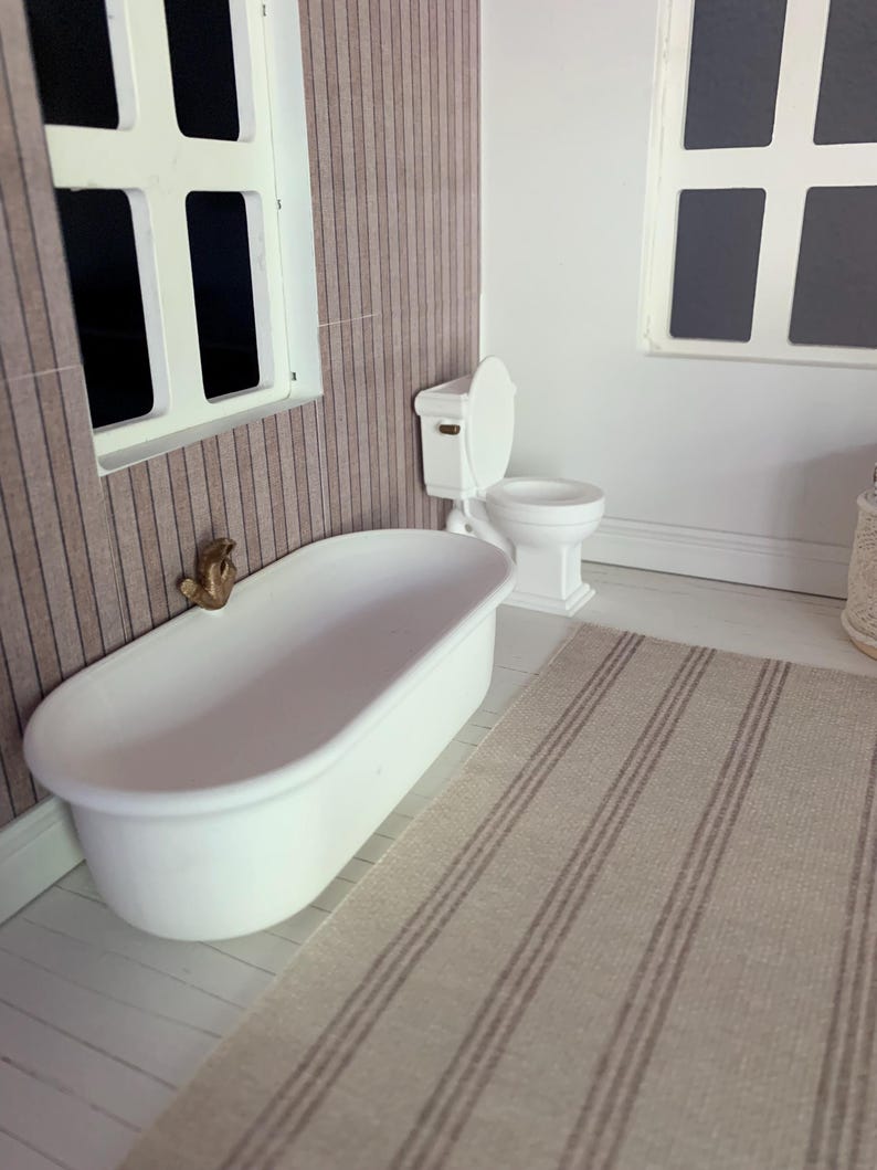 May include: A white bathtub with a gold faucet sits in a bathroom with a white toilet and a striped rug. The bathroom has wood paneling on the walls.
