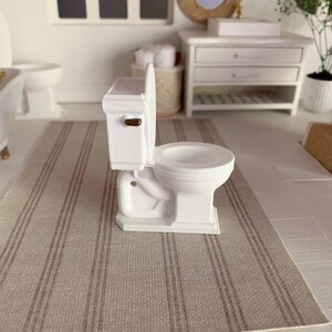 May include: White miniature toilet with a gold handle, sitting on a striped rug in a dollhouse bathroom.