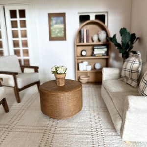 Op de afbeelding: Miniatuur poppenhuis woonkamer sc&egrave;ne met een lichtgekleurde bank, fauteuil en een ronde houten salontafel. Een klein tapijt met een ruitpatroon bedekt de vloer. Een kleine boekenkast en een ingelijste foto zijn zichtbaar.