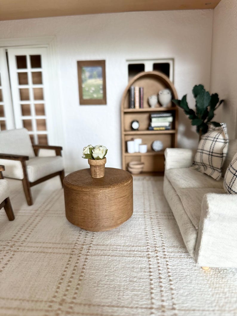 Op de afbeelding: Miniatuur poppenhuis woonkamer sc&egrave;ne met een lichtgekleurde bank, fauteuil en een ronde houten salontafel. Een klein tapijt met een ruitpatroon bedekt de vloer. Een kleine boekenkast en een ingelijste foto zijn zichtbaar.