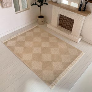 May include: A beige and white diamond patterned rug with a fringed edge. The rug is laid out on a white wood floor in a dollhouse setting.